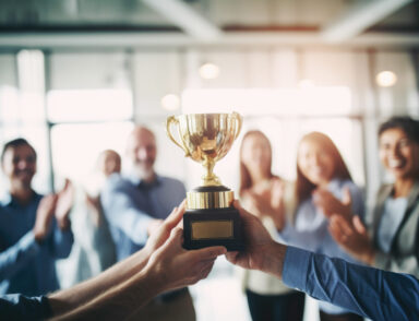 L'image montre deux mains tenant un trophée doré au premier plan, symbolisant une réussite collective ou une victoire.